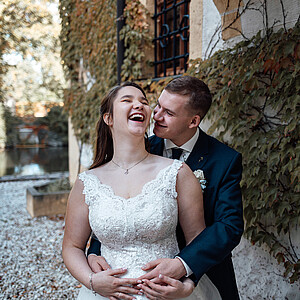 Ein Brautpaar teilt einen verspielten Moment an seinem Hochzeitstag. Die Braut lacht ausgelassen, während der Bräutigam sie umarmt und ihr verschmitzt zulächelt. Im Hintergrund eine mit Efeu bewachsene Wand in warmen Herbstfarben.