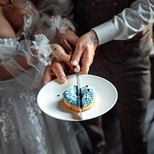 Braut und Bräutigam schneiden gemeinsam einen herzförmigen Donut mit blauer Glasur an, ihre Hände sind dabei ineinander verschränkt.