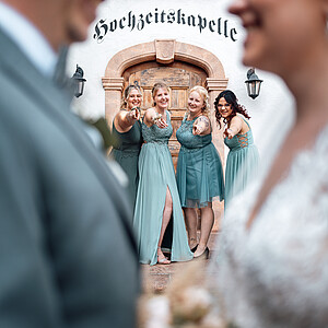 Ein verspieltes Hochzeitsfoto mit der Braut und dem Bräutigam unscharf im Vordergrund. Im Hintergrund stehen vier Brautjungfern in passenden türkisfarbenen Kleidern, die lachend in die Kamera zeigen. Sie posieren vor einer Kapelle mit der Aufschrift „Hochzeitskapelle“.