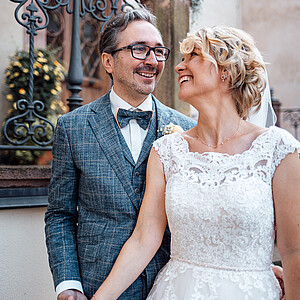 Eine Braut und ein Bräutigam stehen nah beieinander und lachen sich glücklich an. Der Bräutigam, mit Brille und kariertem Anzug, hält die Hand seiner Braut, die in einem eleganten Spitzenkleid strahlt. Im Hintergrund sind kunstvolle Eisenverzierungen und eine steinerne Treppe zu sehen.