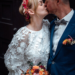 [Romantische Nahaufnahme] Ein frisch verheiratetes Paar teilt einen zärtlichen Kuss vor einer dunklen Holztür. Die Braut trägt ein weißes Spitzenkleid mit Blumenmuster, einen pink-roten Blumenkranz und hält einen bunten Blumenstrauß. Der Bräutigam, gekleidet in einen blauen Anzug mit Blumendeko am Revers, lehnt sich für den Kuss vor.