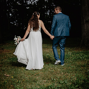 Die Braut und der Bräutigam gehen Hand in Hand durch eine Waldlichtung, von hinten aufgenommen. Das lange weiße Kleid der Braut schwingt im Gehen, während sie ihren Blumenstrauß trägt. Der Bräutigam in seinem petrolfarbenen Anzug schaut lächelnd zu ihr zurück.