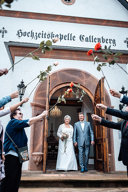 Ein frisch verheiratetes Paar verlässt Hand in Hand die historische Kapelle 'Hochzeitskapelle Callenberg'. Sie lächeln, während sie durch ein Spalier aus Rosen schreiten, das von ihren Gästen gehalten wird. Die Braut trägt ein weißes Spitzenkleid und hält einen pastellfarbenen Blumenstrauß, während der Bräutigam einen silber-blauen Anzug mit passender Fliege trägt."