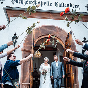 Ein frisch verheiratetes Paar verlässt Hand in Hand die historische Kapelle 'Hochzeitskapelle Callenberg'. Sie lächeln, während sie durch ein Spalier aus Rosen schreiten, das von ihren Gästen gehalten wird. Die Braut trägt ein weißes Spitzenkleid und hält einen pastellfarbenen Blumenstrauß, während der Bräutigam einen silber-blauen Anzug mit passender Fliege trägt."