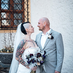 Eine Braut mit halb rasierten Haaren und kunstvollen Tattoos schaut ihrem Bräutigam liebevoll in die Augen. Sie hält einen Brautstrauß in tiefem Lila und Weiß, während er eine passende Ansteckblume zu seinem grau karierten Anzug trägt. Sie stehen vor einem rustikalen Gebäude mit einem Fenster, das von dekorativen Eisenstäben umrahmt ist.