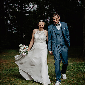 Ein frisch vermähltes Paar geht Hand in Hand über eine Wiese, umgeben von Bäumen. Die Braut trägt ein fließendes weißes Kleid mit Spitzenverzierung und hält einen Blumenstrauß, während der Bräutigam einen petrolfarbenen Anzug mit Fliege und weiße Sneaker trägt. Beide lächeln glücklich.