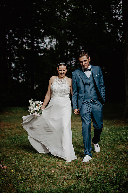 Ein frisch vermähltes Paar geht Hand in Hand über eine Wiese, umgeben von Bäumen. Die Braut trägt ein fließendes weißes Kleid mit Spitzenverzierung und hält einen Blumenstrauß, während der Bräutigam einen petrolfarbenen Anzug mit Fliege und weiße Sneaker trägt. Beide lächeln glücklich.