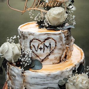 Eine rustikale, zweistöckige Hochzeitstorte mit Birkenholz-Optik, dekoriert mit weißen Rosen und grünen Blättern. Oben steckt ein Holzschriftzug mit den Namen „Vanessa & Martin“, in der Mitte ist ein Herz mit den Initialen „V+M“ in die Glasur eingearbeitet.