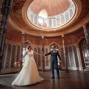 Ein frisch verheiratetes Paar tanzt in einem prunkvollen, runden Saal mit kunstvollen Verzierungen. Die Braut trägt ein fließendes weißes Kleid und hält einen Blumenstrauß, während der Bräutigam in einem karierten Anzug sanft nach ihrer Hand greift. Die hohe Kuppeldecke und filigranen Muster verleihen dem Moment eine märchenhafte Atmosphäre.