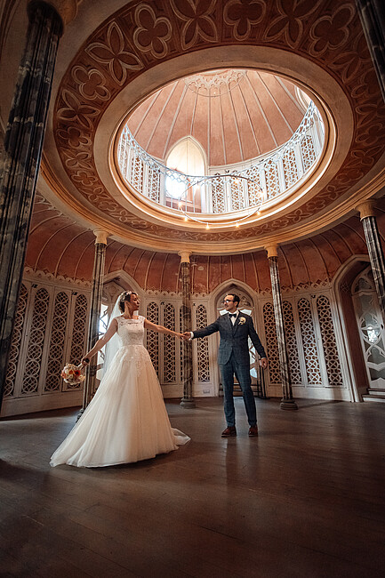 Ein frisch verheiratetes Paar tanzt in einem prunkvollen, runden Saal mit kunstvollen Verzierungen. Die Braut trägt ein fließendes weißes Kleid und hält einen Blumenstrauß, während der Bräutigam in einem karierten Anzug sanft nach ihrer Hand greift. Die hohe Kuppeldecke und filigranen Muster verleihen dem Moment eine märchenhafte Atmosphäre.