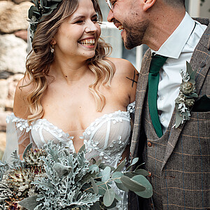 Eine strahlende Braut und ein lachender Bräutigam teilen einen innigen Moment an ihrem Hochzeitstag, beide in rustikaler Kleidung mit grünen Akzenten; die Braut hält einen üppigen, strukturierten Blumenstrauß.