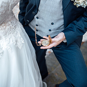 Nahaufnahme eines Bräutigams in einem dunkelblauen Anzug und einer grauen Weste, der eine geöffnete goldene Taschenuhr in der Hand hält. Eine weiße Rosenboutonniere steckt an seinem Revers, und ein Teil des Brautkleids mit Spitze ist neben ihm zu sehen.