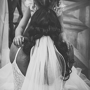 Schwarz-weiß-Fotografie einer Braut, die ein kleines Mädchen in die Arme schließt. Die Braut trägt ein Spitzenkleid mit langem Schleier, während das Mädchen mit geflochtenem Haar und Blumen im Haar ihre Arme um sie legt. Eine innige und emotionale Umarmung.